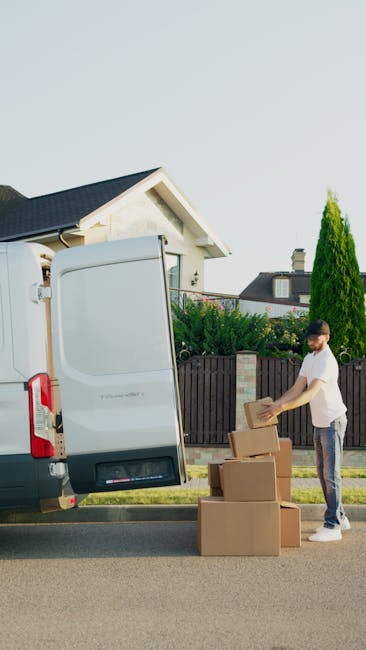 A man dressed in a black vest, blue long-sleeved shirt, and black trousers leans against the open rear of a white moving van parked outside a residential property with a brick exterior and a tiled roo