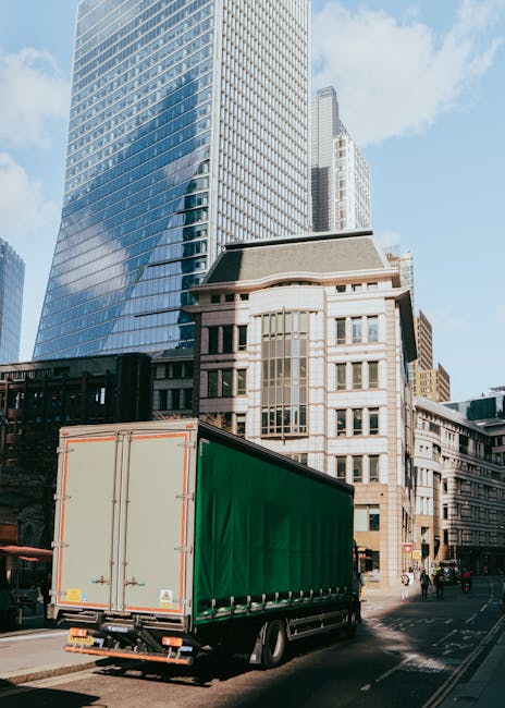 A large green cargo truck with a closed cargo area is parked on a city street next to a sidewalk, with a backdrop of tall modern buildings constructed from glass and concrete. The truck's rear doors are closed, and it appears to be in the process of either loading or unloading, as the vehicle is situated near the curb in an urban environment. The street features a paved road with some pedestrians visible on the sidewalk, and nearby vehicles are parked along the road. The surrounding architecture includes both contemporary high-rise buildings with reflective glass facades and older, more traditional structures with detailed facades. The scene is lit by daylight, indicating clear weather conditions. This image relates to home relocation and furniture transport services provided by Removal Van Lambeth, highlighting the logistics of moving large items in a city setting.