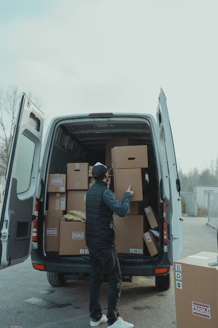 A mover wearing a black jacket, dark pants, and a black cap is inside the open rear of a large white van, which is parked outdoors on a paved surface under an overcast sky. The van's interior is filled with numerous brown cardboard boxes, some stacked on top of each other, each sealed with packing tape and bearing labels. The mover is lifting or adjusting one of the boxes, which is positioned near the doorway of the van. Several other boxes, some on the pavement and others in the loading area, are visible around the vehicle. The van appears ready for home relocation or furniture transport as part of a professional removals service. The background includes a row of trees and a distant industrial or storage building, indicating a typical outdoor setting for a moving process. This scene reflects the packing and loading stages of a house removal, with the focus on securely transporting household items using equipment like packing materials and a large moving van, associated with Removal Van Lambeth's service offerings.
