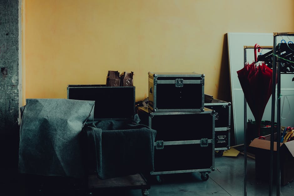 Interior view of a room prepared for a home relocation, showing stacked black equipment cases with metal edges and handles, some on casters, positioned against a yellow wall. To the left, a piece of d