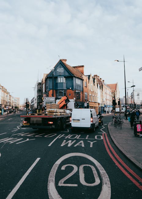 A flatbed removal truck operated by Removal Van Lambeth is parked on a city street in Brixton, preparing for a home relocation. The truck is loaded with wooden furniture pieces, including a pallet and a large, wrapped item, along with cardboard boxes secured on the flatbed. The vehicle is positioned close to the curb, with the loading process in progress, as items are being transferred onto or off the truck. Surrounding the truck are parked bicycles and pedestrians walking along the pavement, which is lined with lamp posts and shopfronts. The street features a clear 20 mph speed limit road marking, with a slight incline leading toward residential buildings in the background, some of which are multi-storey with brick facades and pitched roofs. The scene is illuminated by daylight with a partly cloudy sky overhead. This image exemplifies the logistics involved in furniture transport and packing during a house removals service provided by Removal Van Lambeth, illustrating the typical urban environment for a furniture delivery or home relocation project in Lambeth.