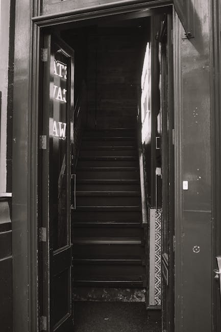 A black-painted staircase leading upward inside a building, viewed through open double doors with glass panels. The staircase has dark wooden steps and a metal handrail on the right side. To the left of the doorframe, reflective signage with partially visible text is mounted on the wall. The surrounding area is illuminated by natural light coming from an overhead source, highlighting the textured wall surfaces and the clean, well-maintained appearance of the staircase. This setting depicts the interior of a property prepared for a home relocation or furniture transport, with the open doors suggesting the beginning of the loading process for a removals service. The image aligns with services offered by Removal Van Lambeth for navigating narrow staircases during house removals, as shown on the webpage 'Clapham Common flat removals tips for narrow staircases' at removalvanlambeth.co.uk.