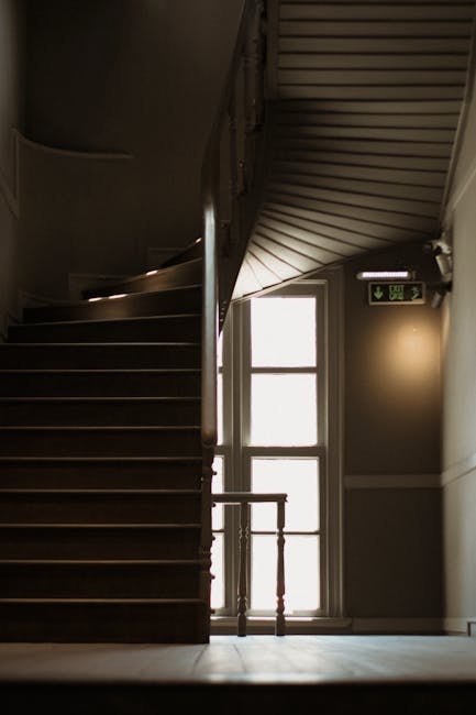 A black-painted staircase leading upward inside a building, viewed through open double doors with glass panels. The staircase has dark wooden steps and a metal handrail on the right side. To the left of the doorframe, reflective signage with partially visible text is mounted on the wall. The surrounding area is illuminated by natural light coming from an overhead source, highlighting the textured wall surfaces and the clean, well-maintained appearance of the staircase. This setting depicts the interior of a property prepared for a home relocation or furniture transport, with the open doors suggesting the beginning of the loading process for a removals service. The image aligns with services offered by Removal Van Lambeth for navigating narrow staircases during house removals, as shown on the webpage 'Clapham Common flat removals tips for narrow staircases' at removalvanlambeth.co.uk.