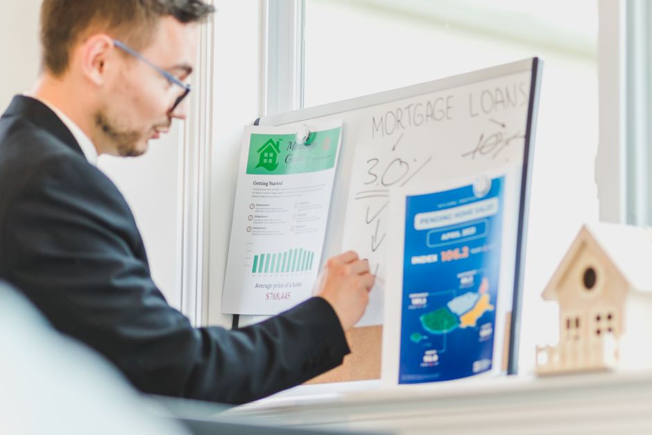 A man in a dark suit and glasses is writing on a whiteboard positioned in an office environment with natural light coming through nearby windows. The whiteboard contains handwritten notes about mortgage loans, including percentages and checkmarks, alongside printed materials clipped to the board, such as a house-related flyer with green and white graphics. To the right of the whiteboard, there is a small wooden model of a house placed on the desk or shelf. The scene suggests planning or discussion related to property or relocation processes, highlighting the professional context connected to house removals and moving services offered by Removal Van Lambeth, especially in regard to commercial office moves in the SE11 area.