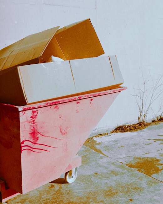 A large pink waste collection skip positioned outdoors on a concrete surface, partially open with a brown cardboard box and what appears to be a white bag inside. The cardboard box is tilted and rests on top of the skip, with its flaps open. Nearby, a small, leafless bush is visible against a plain white wall in the background, indicating an exterior environment. The scene is well-lit with natural daylight, suggesting daytime conditions. The area around the skip is tidy, with some dirt patches and a slight shadow cast by the skip and objects inside. This image reflects the process of waste disposal in a home relocation or moving context, potentially involving the collection of packing materials or discarded items, fitting into services offered by Removal Van Lambeth for efficient and compliant waste removal related to relocations or removals.