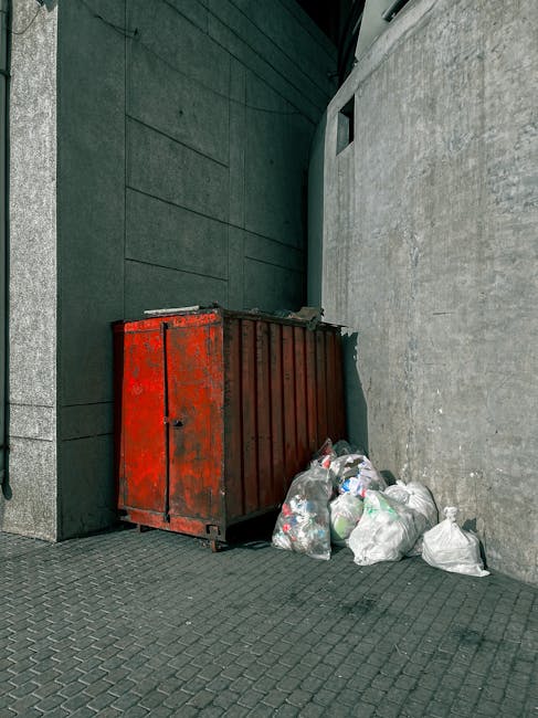 A red metal waste skip with visible rust and chipped paint is positioned against a grey urban wall, near a building with dark green siding. Several white plastic bags filled with assorted rubbish, including paper, plastic bottles, and other household waste, are piled beside the skip on a textured pavement surface. The scene is outdoors, with natural lighting casting soft shadows, indicating daytime. This image illustrates waste collection as part of a home relocation process, which may require removal of bulky waste before moving, a service that companies like Removal Van Lambeth offer in accordance with Lambeth Council rules for skip hire and bulky waste disposal.