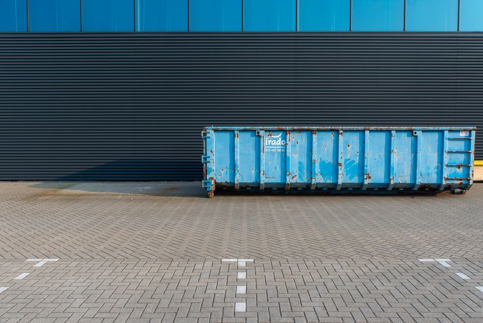A large pink waste collection skip positioned outdoors on a concrete surface, partially open with a brown cardboard box and what appears to be a white bag inside. The cardboard box is tilted and rests on top of the skip, with its flaps open. Nearby, a small, leafless bush is visible against a plain white wall in the background, indicating an exterior environment. The scene is well-lit with natural daylight, suggesting daytime conditions. The area around the skip is tidy, with some dirt patches and a slight shadow cast by the skip and objects inside. This image reflects the process of waste disposal in a home relocation or moving context, potentially involving the collection of packing materials or discarded items, fitting into services offered by Removal Van Lambeth for efficient and compliant waste removal related to relocations or removals.