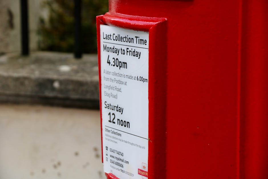 A white paper notice is attached to a red post box on a pavement, displaying scheduled collection times for mail and packages, including last collection from Monday to Friday at 4:30 pm and Saturday at 12 noon. The notice is printed with black text on a white background and has details about collection procedures from the local postbox at Longfield Road (Stag Road), with contact information and reference numbers below. The postbox is situated outdoors next to a building with a concrete doorstep and a metal railing in the background, indicating a typical street scene. The display is illuminated by natural daylight, with a focus on the notice and the red postbox, supporting the understanding of home or community mail collection points often considered during house removals or relocations. The setting relates subtly to logistical considerations involved in moving or delivery processes supported by Removal Van Lambeth's services.