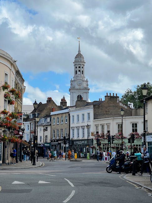 A busy street scene in Lambeth with a prominent white clock tower and spire extending into a partly cloudy sky. The street is lined with multi-story buildings featuring shops with large windows, some with hanging flower baskets and signage. Pedestrians are walking along the pavement, some carrying shopping bags or engaged in conversation. A group of people is gathered near shop entrances, and a couple of scooters are parked on the side of the road. In the foreground, a Loading process for house removals is suggested by the presence of a van partially visible on the right, with the pavement showing signs of current moving activities, including a trolley or packing materials nearby. The overall scene depicts a typical urban area suitable for home relocation and furniture transport services, with visible elements of moving logistics, such as transit vehicles and pedestrian activity, reflecting the ongoing process of packing and moving.