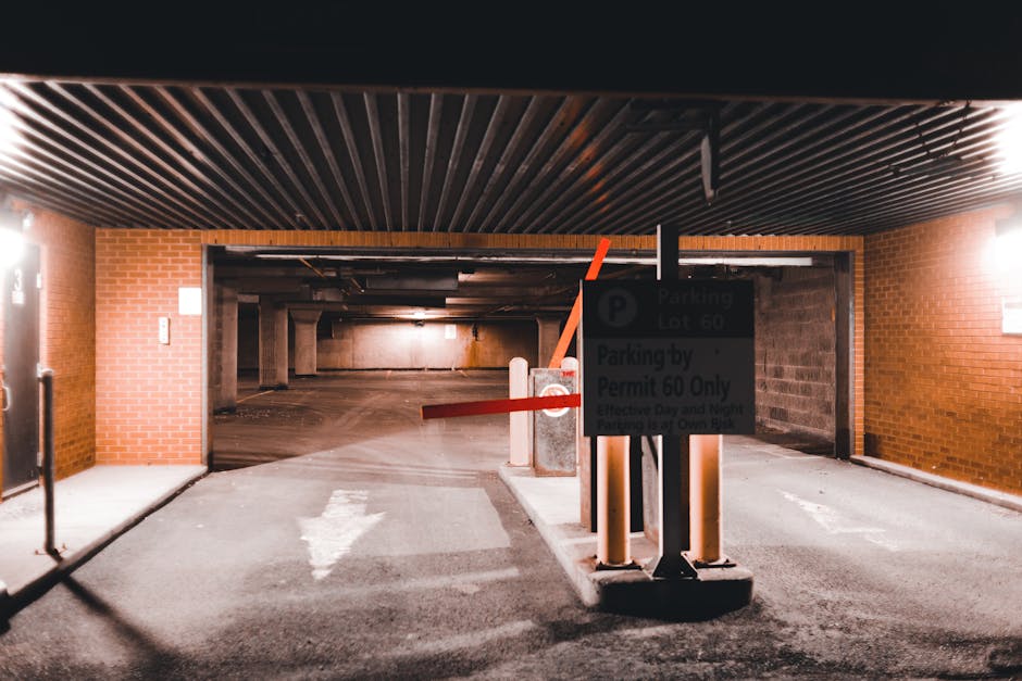 Inside a multi-level parking garage with exposed brick walls and a dark corrugated ceiling, the image shows the entrance to an underground parking area designated for house removals and furniture transport. A large automated barrier arm, painted orange, is raised to permit vehicle access, with a parking signage board fixed to a central pillar in front of the entrance, indicating parking lot information and permit requirements. The pavement features a white directional arrow pointing forward, guiding the loading process through the garage. Light from overhead fixtures illuminates the concrete floor and surrounding brick walls, creating a secure environment for loading and moving furniture and boxes. To the left, part of a door or access point is visible, while the background reveals a spacious, empty parking area with supportive columns and additional vehicle pathways, supporting the logistics for professional home relocation services provided by [COMPANY_NAME].