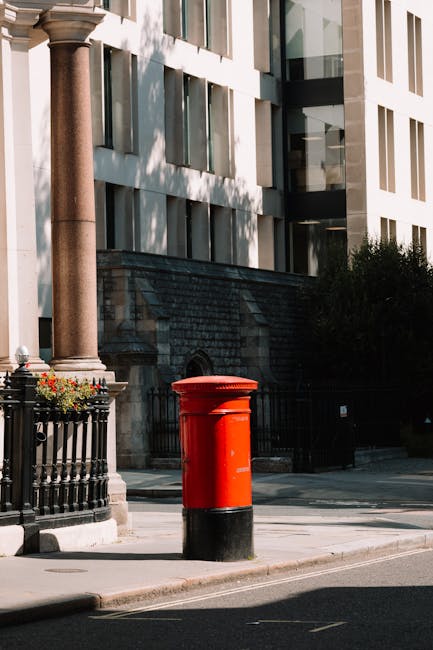 A bright red cylindrical post box with a black base is situated on a sidewalk in front of a modern, multi-story building with large glass windows and white exterior panels. To the left, there is a decorative black metal fence with flower baskets attached, containing blooming red and white flowers. The building's entrance features a stone archway and steps, with a stone pillar next to the post box. The scene appears during daytime with shadows cast by trees and surrounding structures. This setting may relate to residential or commercial property contexts, and [COMPANY_NAME], such as Removal Van Lambeth, provides house removals or furniture transport services in the area, supporting home relocation projects and moving logistics.
