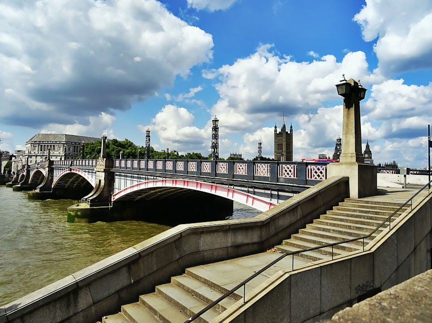 A daytime view of Westminster Bridge spanning the River Thames in London, with the iconic Houses of Parliament and Big Ben visible in the background under a blue sky with scattered white clouds. In the foreground, stone stairs with black metal handrails lead up from a paved walkway along the riverbank, which is partially obscured by the bridge. The scene is well-lit with natural sunlight, casting shadows on the stone structures. This location exemplifies urban riverfront scenery often seen during home relocations or moving logistics situated near key London landmarks, supported by companies such as Removal Van Lambeth, who assist with furniture transport and packing during house removals.