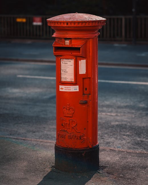 A bright red cylindrical post box with a black base is situated on a sidewalk in front of a modern, multi-story building with large glass windows and white exterior panels. To the left, there is a decorative black metal fence with flower baskets attached, containing blooming red and white flowers. The building's entrance features a stone archway and steps, with a stone pillar next to the post box. The scene appears during daytime with shadows cast by trees and surrounding structures. This setting may relate to residential or commercial property contexts, and [COMPANY_NAME], such as Removal Van Lambeth, provides house removals or furniture transport services in the area, supporting home relocation projects and moving logistics.