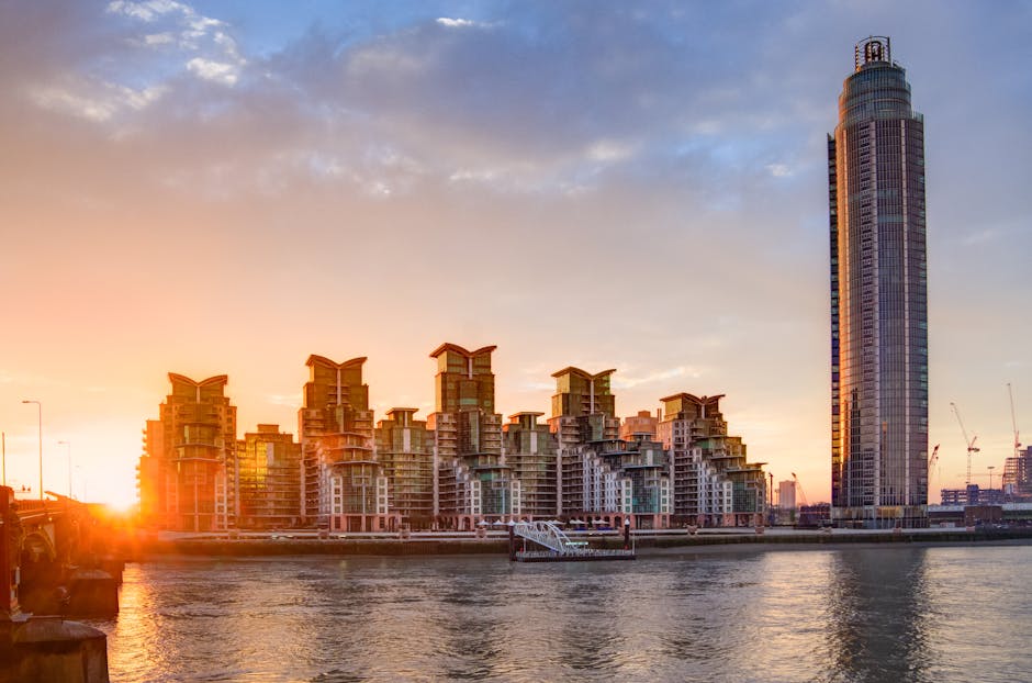 A city skyline at sunset featuring a tall, cylindrical glass skyscraper on the right and a cluster of modern, multi-storey residential buildings with distinctive rooftop structures on the left, situated along a waterway. The buildings are under construction with cranes visible in the background. In the foreground, a section of a residential building's doorway is visible, showing a wooden doorframe and a glimpse of an interior, with a few cardboard boxes and packing materials on the pavement near the entrance. A moving van from Removal Van Lambeth is parked nearby, with a pallet jack and moving blankets in the loading area. The scene captures the ongoing home relocation process through the loading and unloading of furniture and packing boxes as part of a furniture transport and packing and moving service. The sky is partly cloudy, illuminated by the warm hues of the setting sun, creating a contrast between the activity of moving logistics and the serene urban waterway environment.