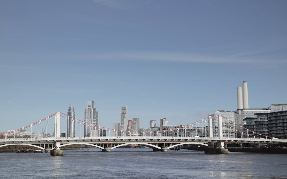 A wide-angle view of a white suspension bridge spanning a river with visible cables and towers, connecting two parts of a modern city skyline in the background. Behind the bridge, there are high-rise buildings made of glass and steel, including a tall tower with a pointed top, and several other skyscrapers with varying heights and architectural styles. On the right side of the image, there are industrial buildings with two tall white chimneys, and a large, multi-story structure with balconies. The scene is captured during daylight with clear blue skies, highlighting the urban landscape. The river below shows reflections of the bridge and city, and the scene depicts a typical urban environment suitable for house removals, involving the transport of furniture and boxes across the city, possibly by a professional removal service like Removal Van Lambeth.